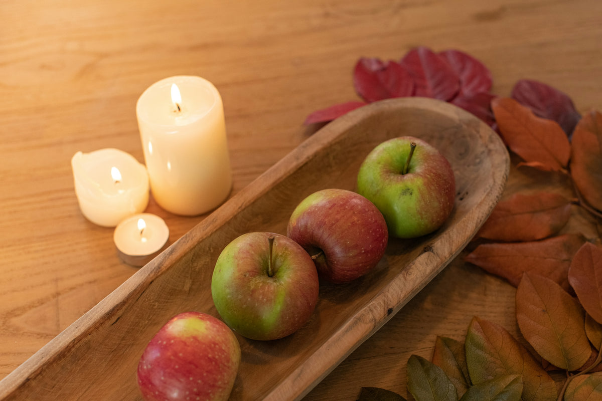 Brandende witte stompkaarsen en waxinelichtjes naast een houten schaal met appels en herfstbladeren