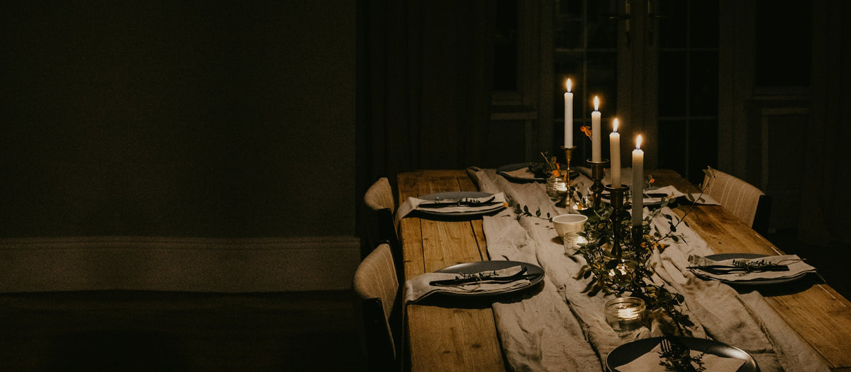 Lange houten tafel met linnen loper, gedekte borden en brandende witte dinerkaarsen in koperen kandelaars